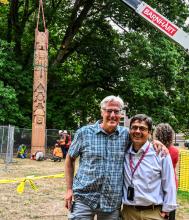 Two individuals stand in the foreground smiling as the Coast Salish Story Pole is raised vertically by a crane