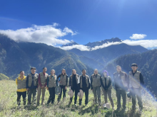 A group of individuals stand in the foreground high on top of a mountain with several mountain tops and clouds behind them