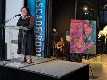 Ronalei stands behind a podium on a stage speaking to the audience, wearing a black dress and has a pinkish red flower in their hair