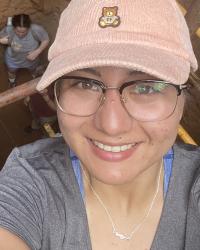 Woman with glasses in a pink baseball hat with a teddy bear on it, standing in a dirt hole 