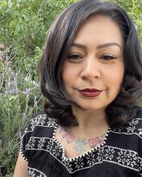 Image is of an Indigenous woman wearing a traditional hand-stitched top who is smiling in front of an elderberry brush