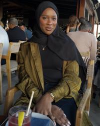 Anaan smiles at the camera with her hands crossed on her lap, wearing a black hijab, black shirt, and olive green jacket