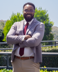 Julius, wearing a gray jacket over a white button down shirt and red tie, smiles with his arms crossed