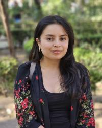 Darkhaired woman in floral overshirt with background of bushes.