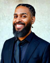 Image of Jamaal Muwwakkil in a black suit against a blurred white background