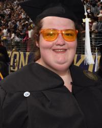 A woman smiles at the camera while wearing orange framed glasses, a graduation hat, and a graduation gown