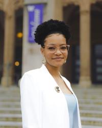 Kennedy, wearing a white blazer over a light-blue undershirt and glasses, smiles while standing in front of a building