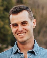 White man with short brown hair and brown eyes in a denim button down shirt in front of some trees.