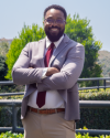 Julius, wearing a gray jacket over a white button down shirt and red tie, smiles with his arms crossed