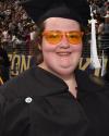 A woman smiles at the camera while wearing orange framed glasses, a graduation hat, and a graduation gown