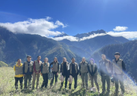 A group of individuals stand in the foreground high on top of a mountain with several mountain tops and clouds behind them