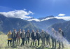 A group of individuals stand in the foreground high on top of a mountain with several mountain tops and clouds behind them